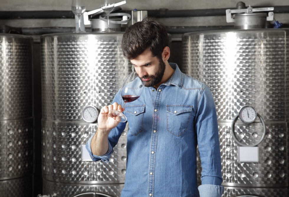 Portrait of winery owner at work. Young professional winemaker standing in front of stainless steel vessel while tasting a glass of red wine.