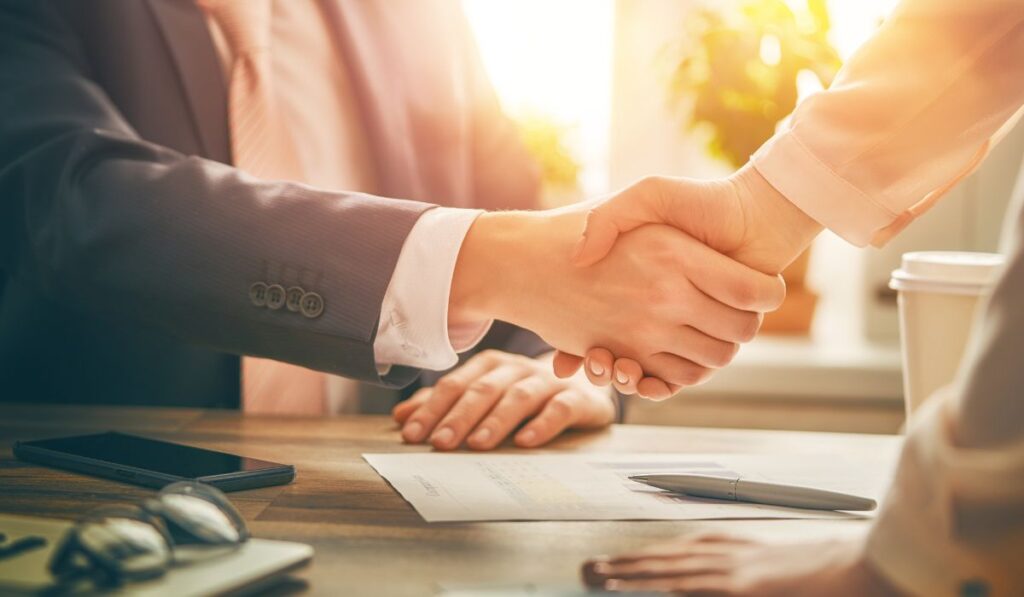 Protea Financial Communication with Financial Professionals man and woman shaking hands over a desk with paperwork, a cell phone, and glasses on the desk
